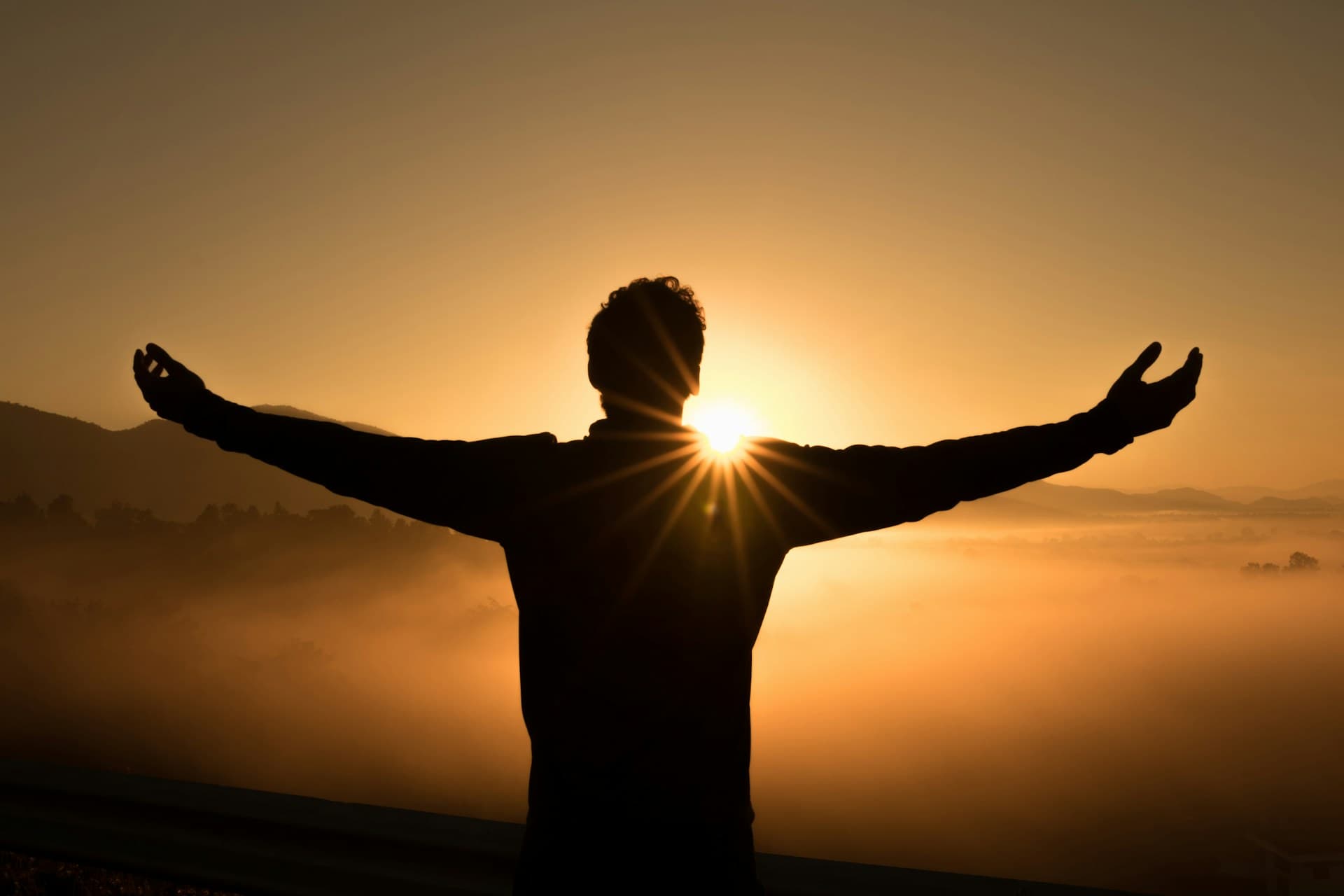 Silhouette of a person on a hill at sunset with arms raised toward warm light.
